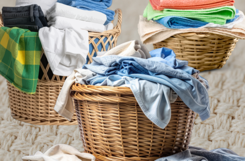An image of three large woven laundry baskets packed with clean, folded clothes and towels. Bright greens, blues, and whites are visible. A few items of clothing lie softly on the plush rug in the foreground. The background is a neutral, textured wall.