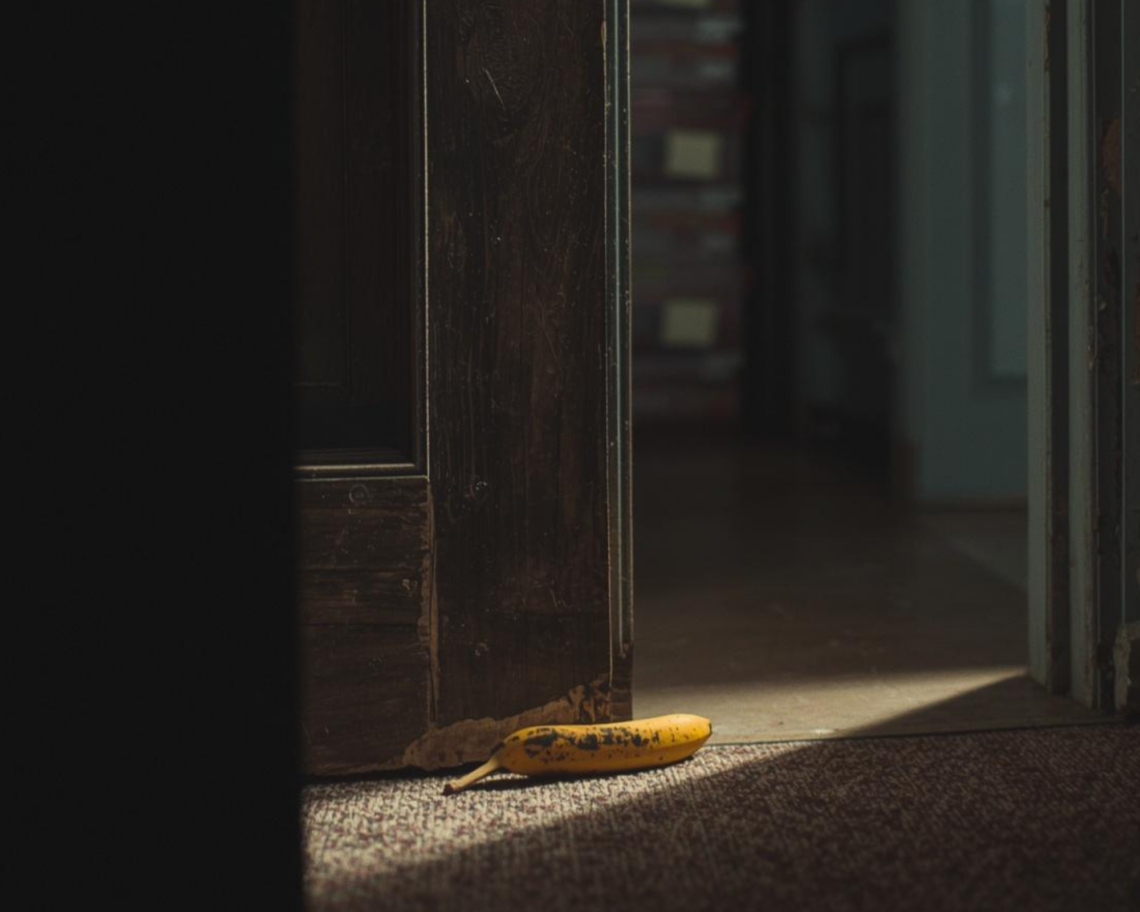 A bright yellow banana with small brown spots lies on a dark textured carpet next to a vintage dark-brown wooden door frame. The door is slightly open, revealing a glimpse of a shadowed hallway beyond. A dramatic shaft of light illuminates the fruit on the floor