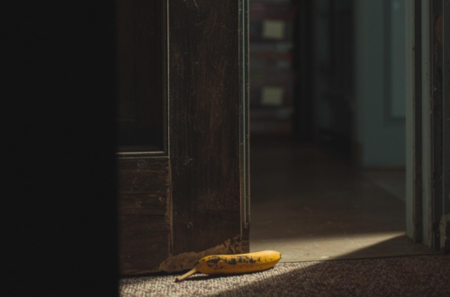 A bright yellow banana with small brown spots lies on a dark textured carpet next to a vintage dark-brown wooden door frame. The door is slightly open, revealing a glimpse of a shadowed hallway beyond. A dramatic shaft of light illuminates the fruit on the floor