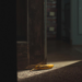 A bright yellow banana with small brown spots lies on a dark textured carpet next to a vintage dark-brown wooden door frame. The door is slightly open, revealing a glimpse of a shadowed hallway beyond. A dramatic shaft of light illuminates the fruit on the floor