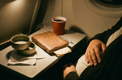 A close-up shot of a fold-down airplane tray table in a dimly lit cabin. Two empty metallic tuna tins are stacked on a white napkin. A brown leather journal with a pen and a red paper cup are nearby. To the right, a passenger in a dark sweater rests their hand on their belly. The scene is illuminated by a warm overhead reading light.
