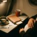 A close-up shot of a fold-down airplane tray table in a dimly lit cabin. Two empty metallic tuna tins are stacked on a white napkin. A brown leather journal with a pen and a red paper cup are nearby. To the right, a passenger in a dark sweater rests their hand on their belly. The scene is illuminated by a warm overhead reading light.