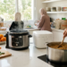 A bright, airy kitchen scene showing the faceless hands of a Muslim mother and her children preparing a large meal. In the foreground, a mother's hands stir a steaming stainless steel pot on a modern stovetop. Nearby, children's hands are busy chopping vegetables and reaching for fresh fruit. On the side counter, a multi-cooker and rice cooker sit among grocery bags, while a hand-designed family chore chart is visible on the wall in the background. The atmosphere is warm, sun-drenched, and captures the busy, organized rhythm of a large household