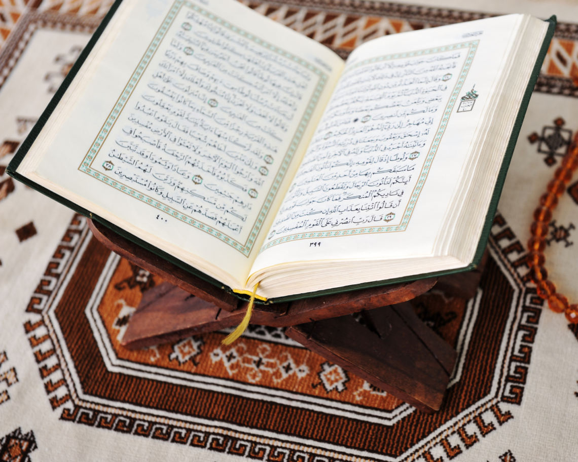 A close-up, light and airy shot of a woman’s hands holding an open Quran. Soft morning sunlight illuminates the pages and a simple notebook and pen resting nearby. The scene is faceless and serene, focusing on the texture of the paper and the quiet moment of reflection.