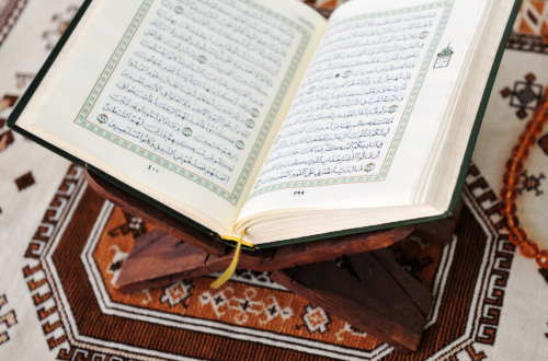 A close-up, light and airy shot of a woman’s hands holding an open Quran. Soft morning sunlight illuminates the pages and a simple notebook and pen resting nearby. The scene is faceless and serene, focusing on the texture of the paper and the quiet moment of reflection.