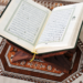 A close-up, light and airy shot of a woman’s hands holding an open Quran. Soft morning sunlight illuminates the pages and a simple notebook and pen resting nearby. The scene is faceless and serene, focusing on the texture of the paper and the quiet moment of reflection.