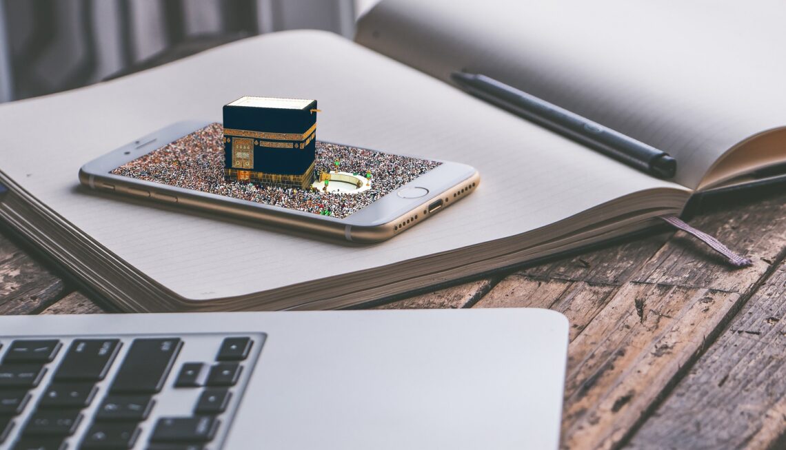 An open notebook rests on a rustic wooden table, its blank lined pages waiting to be filled. On top of the notebook lies a silver smartphone, its screen displaying a digital composite image: a miniature 3D model of the Kaaba, surrounded by a dense crowd of pilgrims gathered in the courtyard of the Masjid al-Haram. A black stylus pen rests in the crease of the open journal. In the foreground, the silver edge and black keys of a laptop keyboard are softly blurred, completing a quiet workspace setting that merges analog reflection with digital connection.