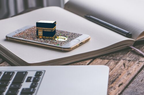 An open notebook rests on a rustic wooden table, its blank lined pages waiting to be filled. On top of the notebook lies a silver smartphone, its screen displaying a digital composite image: a miniature 3D model of the Kaaba, surrounded by a dense crowd of pilgrims gathered in the courtyard of the Masjid al-Haram. A black stylus pen rests in the crease of the open journal. In the foreground, the silver edge and black keys of a laptop keyboard are softly blurred, completing a quiet workspace setting that merges analog reflection with digital connection.