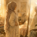 A soft, warm watercolor illustration in a gentle children's book aesthetic showing a Muslim mother in a long cream-colored gown and a dusty rose hijab. She stands in side profile, looking thoughtfully into a large full-length mirror in a bedroom filled with amber afternoon sunlight. Her hand rests gently on her waist as she contemplates her reflection. In the background, elements of a full, busy life are visible: a neatly folded stack of laundry on a chair, a children's book titled "The Little Muslimah" on a nightstand, and a framed Quranic reminder hanging on the wall that reads "And rely upon Allah; and sufficient is Allah as Disposer of affairs (33:3)." Subtle, magical light effects and star dust trail softly across the foreground.